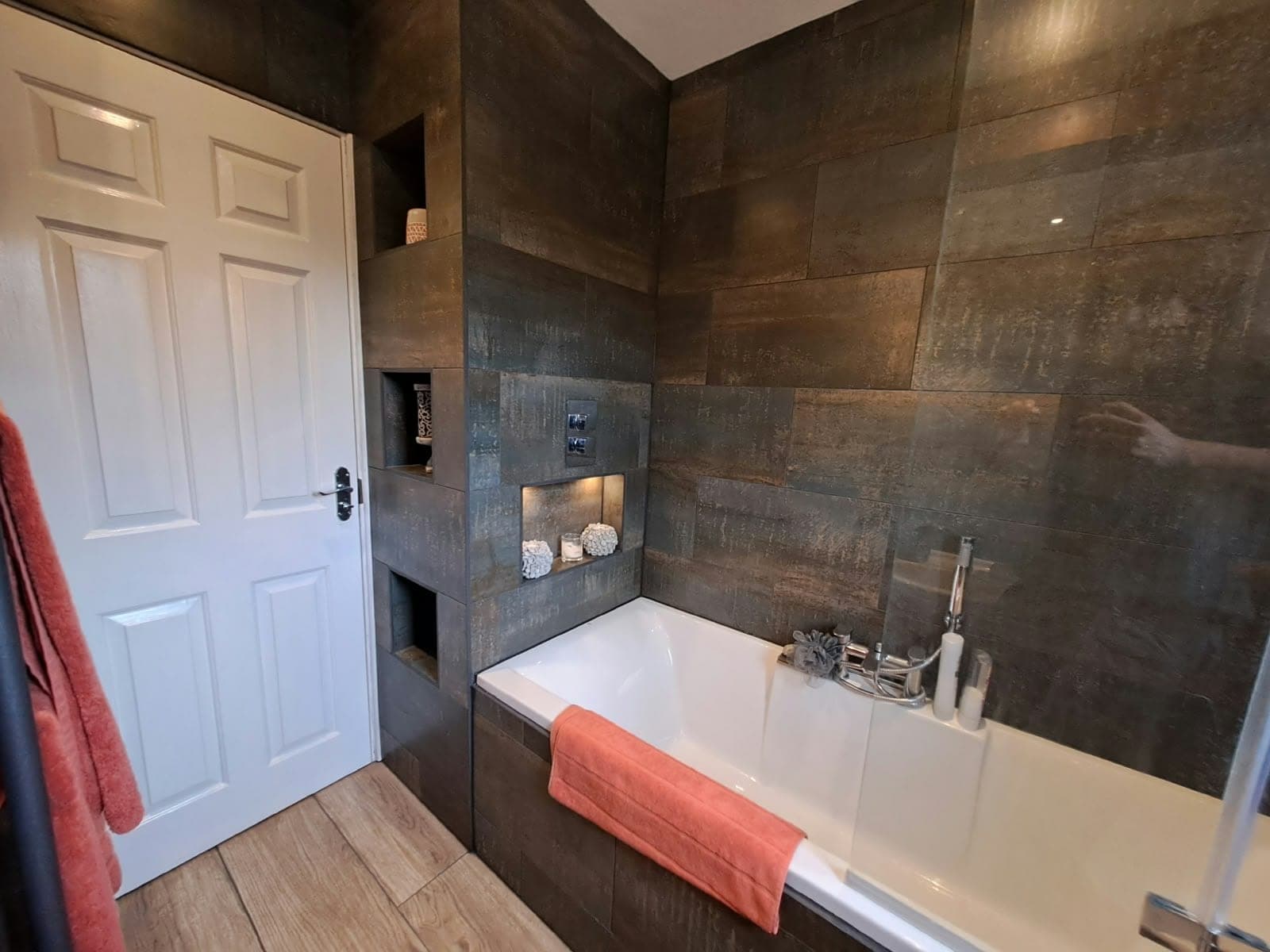 Bathroom with dark metallic-effect wall tiles, illuminated recessed niches and a white bath with a shower screen