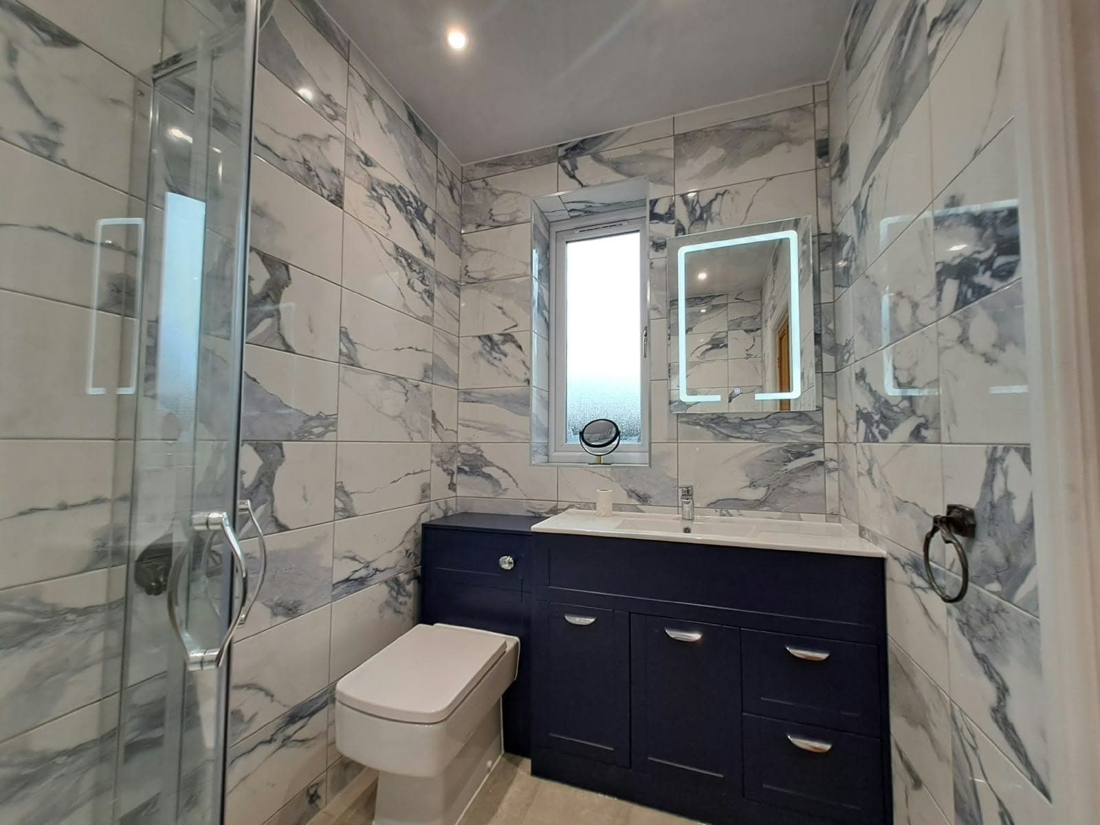 En-suite with marble-effect wall tiles, a navy fitted vanity unit, back-to-wall toilet and illuminated mirror