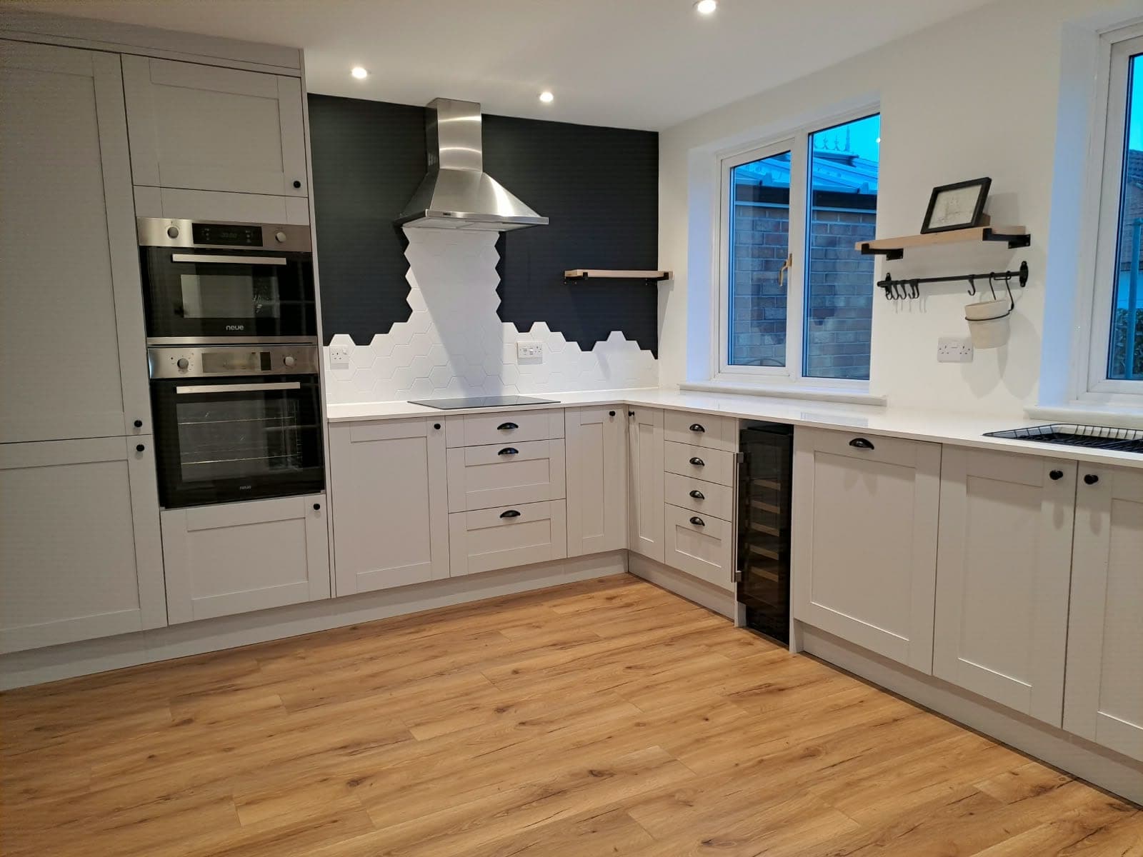 Light grey Shaker-style kitchen with white worktops, built-in double oven and white hexagon tiles against a dark feature wall
